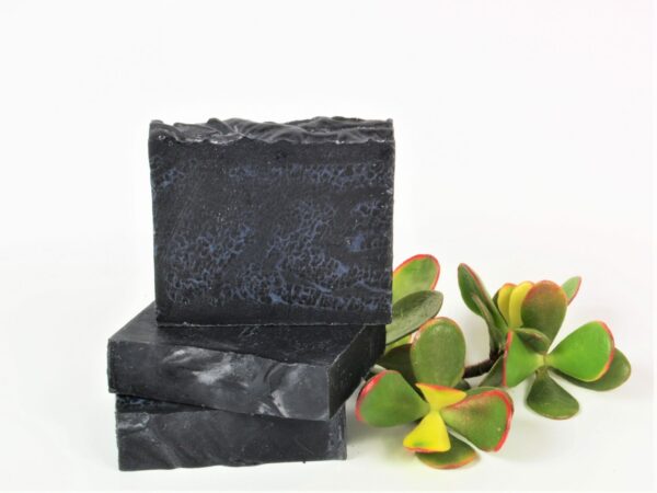three charcoal and rose geranium soaps in a stack next to plant on a white background